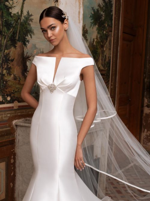 Model wears a Fitted Mikado Wedding Dress called Altair by Bridal Gown Designer Pronovias. The Bardot neckline draws your eye to the slight plunge and the spectacular brooch. Surper sexy
